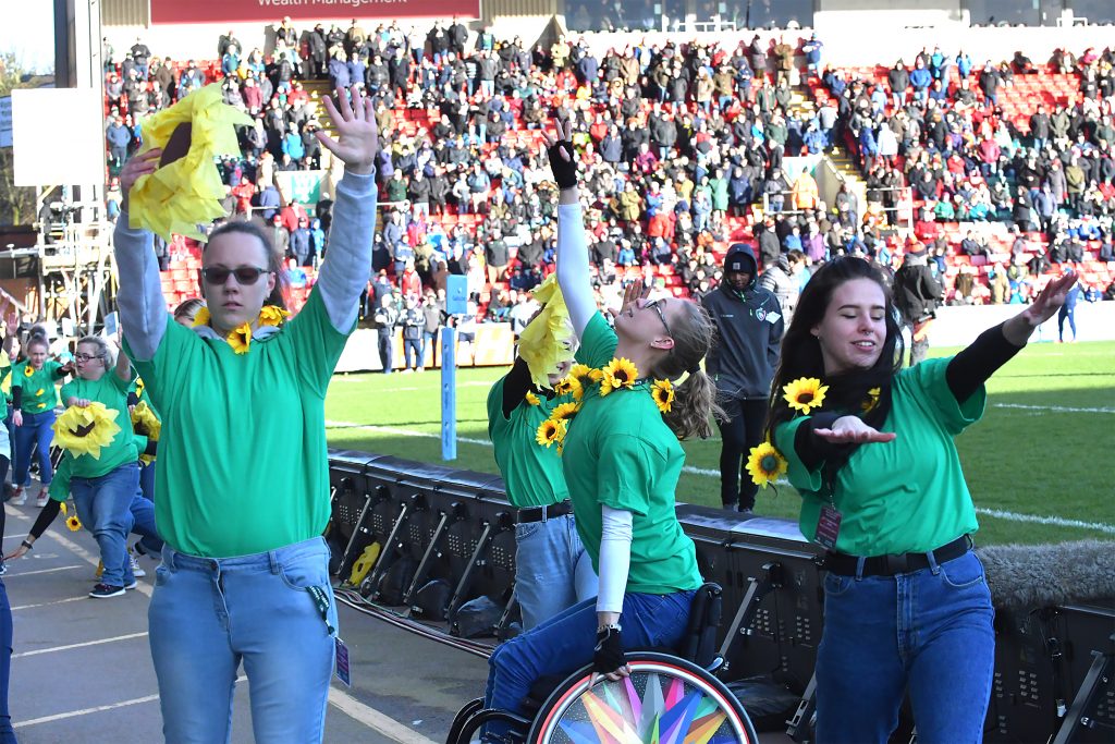 Dancers in green t-shirts holding sunflowers, in front of the crowd at Welford Road