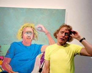 Grayson Perry standing, posing in front of his own portrait by artist Lucy Jones.