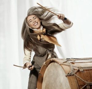 Dame Evelyn Glennie holding a stick in each hand about to hit a large drum with her hair in motion with a big smile on her face