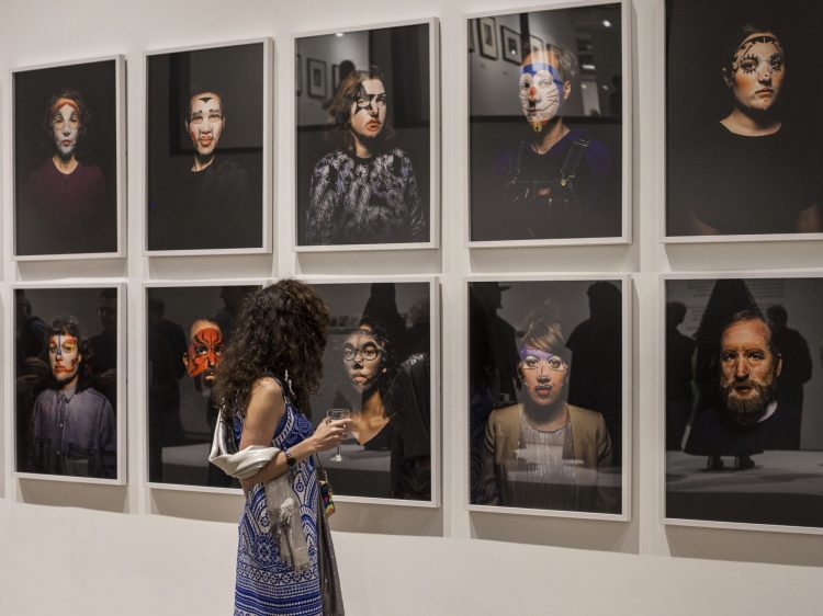 A woman with long dark hair standing in front of a wall of portrait photographs where each person wears full-face makeup.