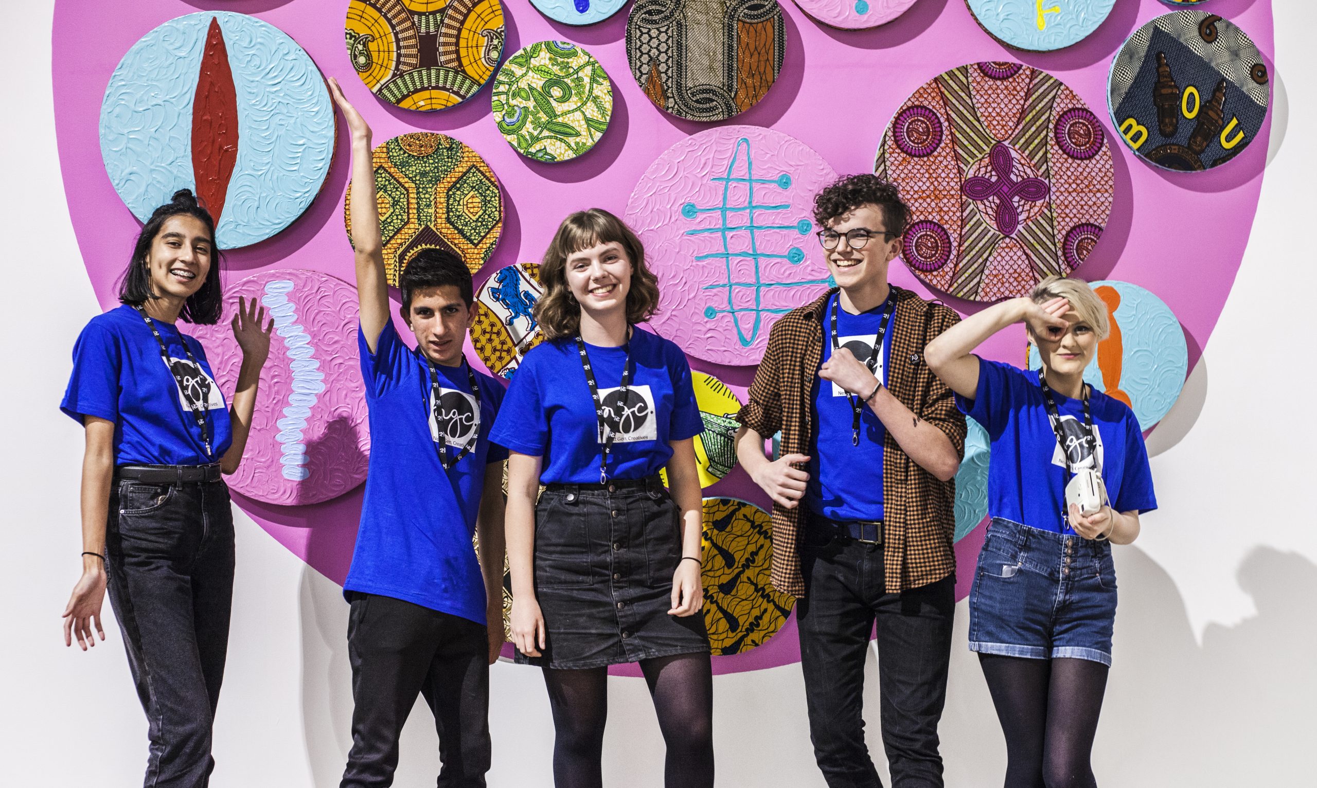 Five smiling people in blue t-shirts posing in front of a pink circle of artworks on a white wall.