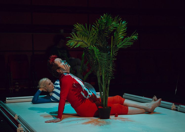 a woman wearing a red suit, sits by a potted plant, on a modelling runway