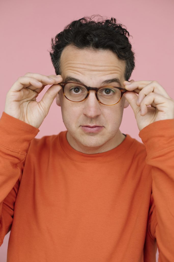 A man with short dark curly hair wearing an orange jumper posing holding his glasses on his face.