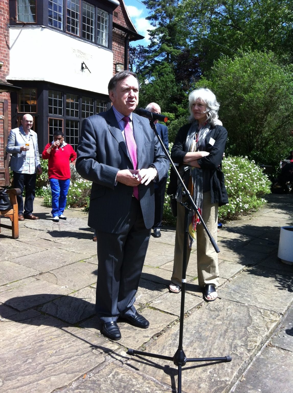 A man with grey-white hair wearing a navy suit giving a speech in the Botanical Gardens.