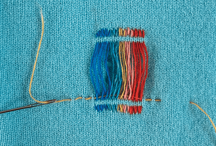 A knit jumper that is being mended with rainbow threads.