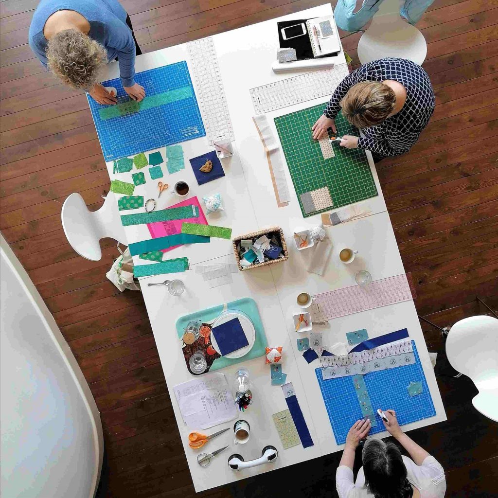 A top-down view of patchwork supplies laid out on a table and three people working at the table.