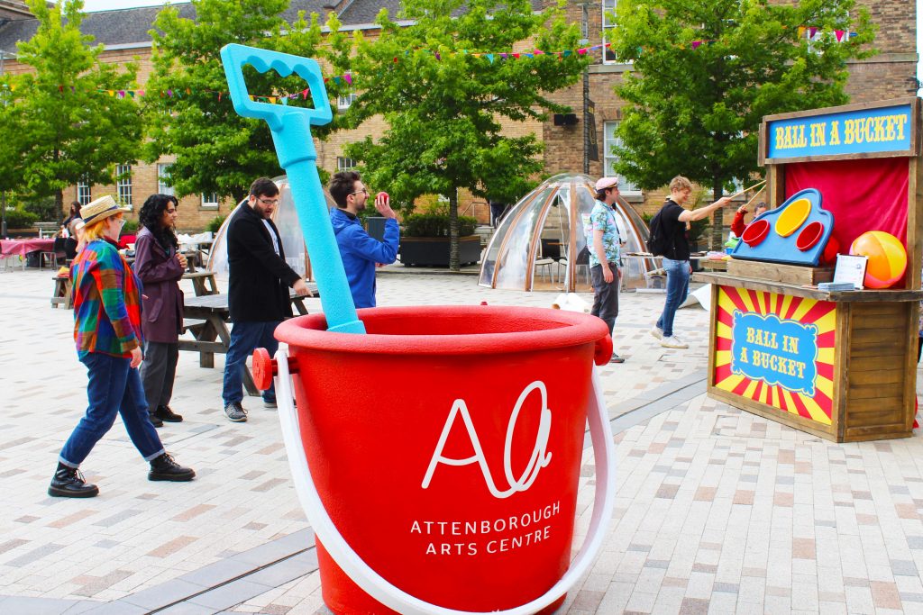 A huge red bucket with the AAC logo on it and a blue spade inside sit in the foreground, while in the background people play Ball in a Bucket.