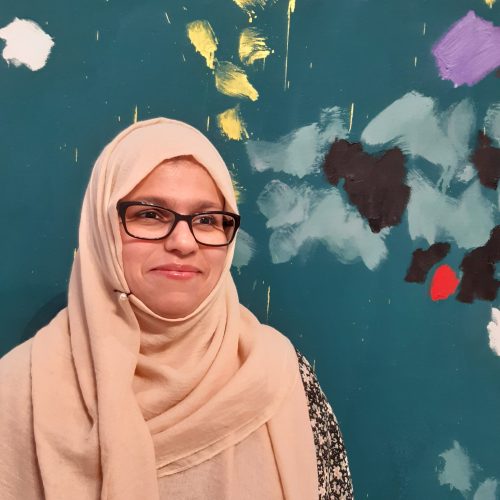 A woman wearing a peach hijab and glasses standing in front of a painted background and smiling to the side of the camera.