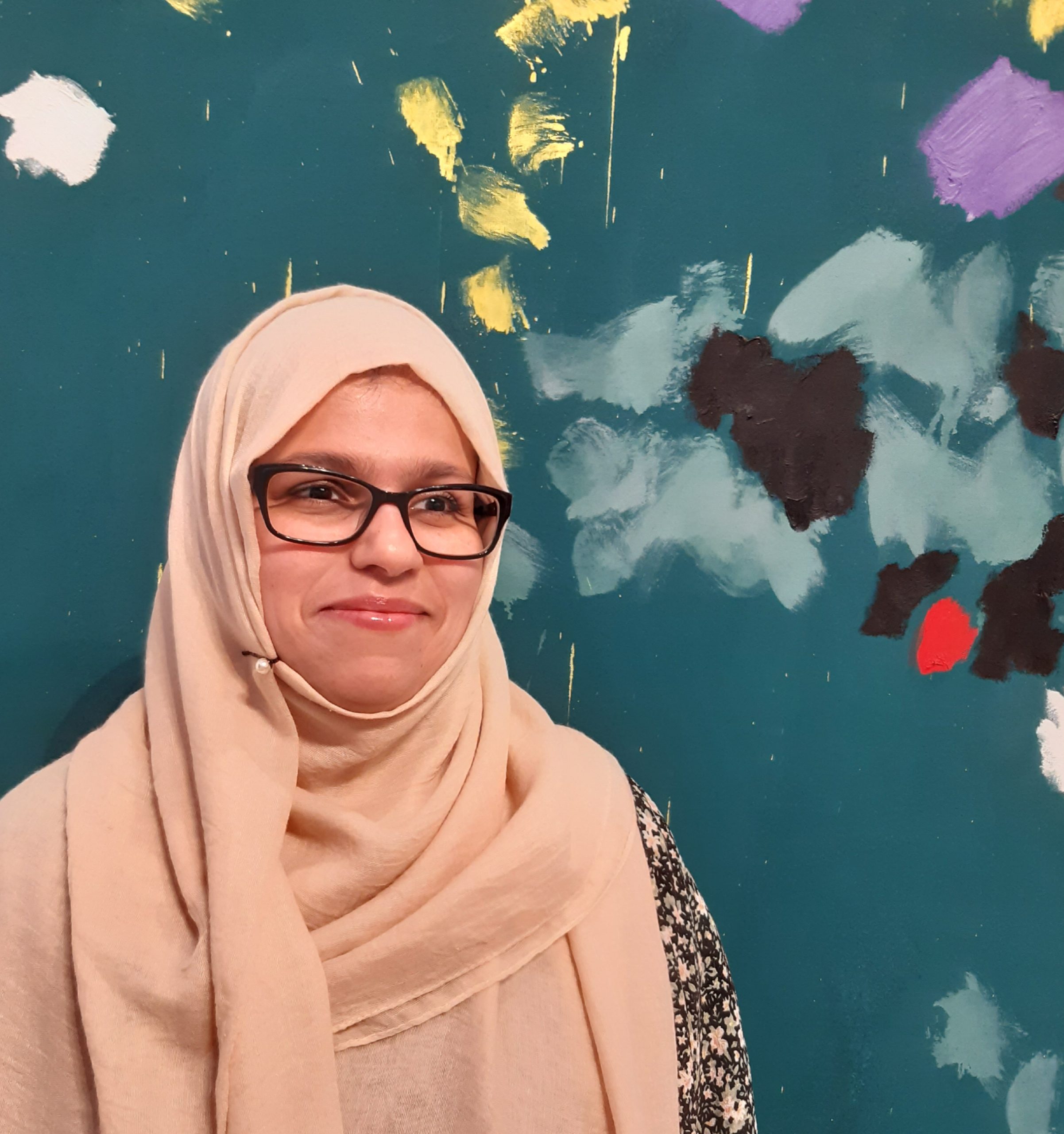 A woman wearing a peach hijab and glasses standing in front of a painted background and smiling to the side of the camera.