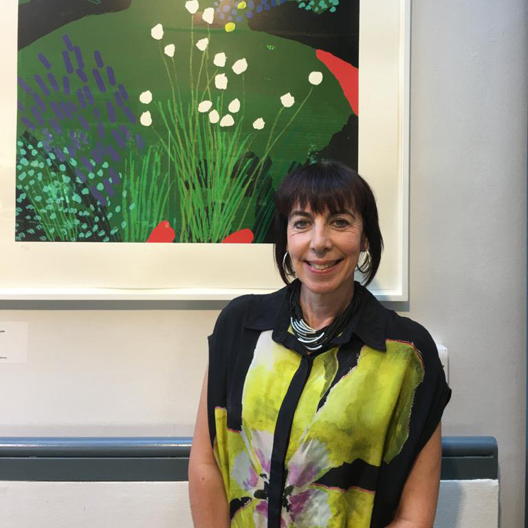 A woman with short, black hair with a fringe smiling at the camera and standing in front of a framed artwork.