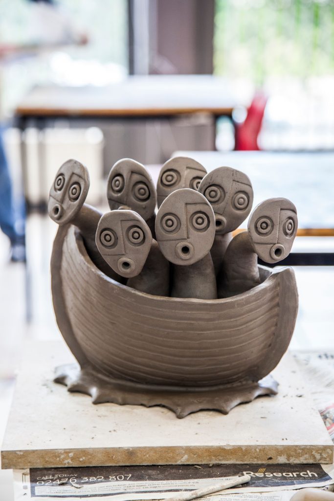 Image of clay sculpture of a boat with 8 heads poking out. Each face has an 'ooo' facial expression.