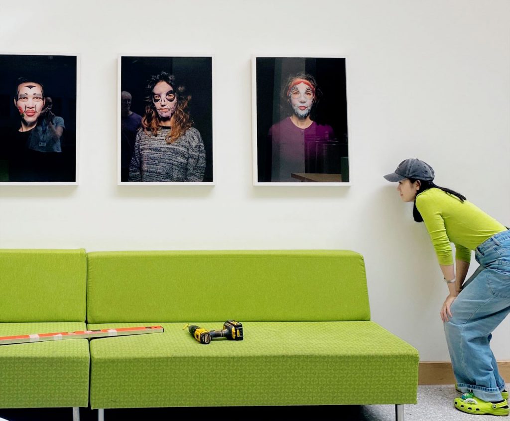 Student looks to the left of a series of framed photographs to check they are level. She is wearing a matching green to the sofa which is under the framed pieces.