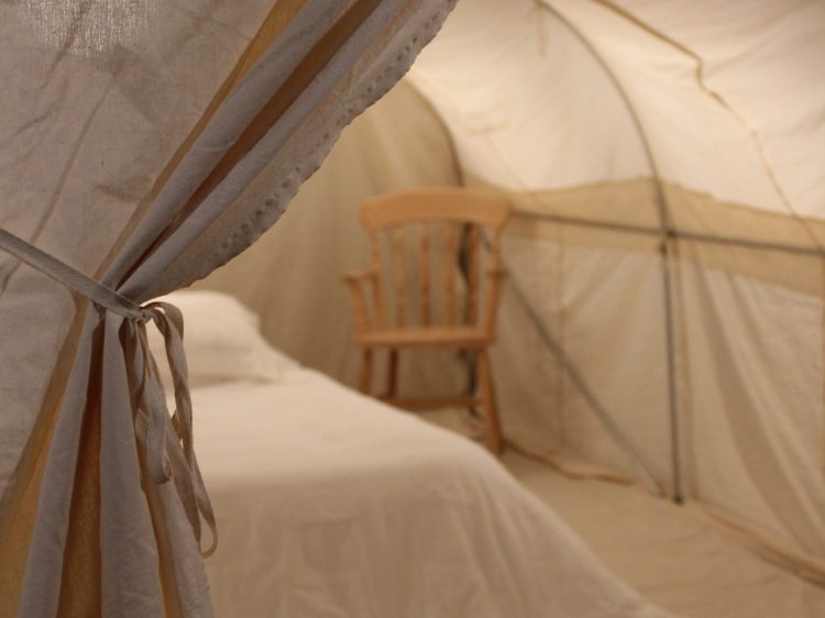 A white curtain pulled back to show the inside of a canvas, cream tent, with a white bed and wooden chair inside.