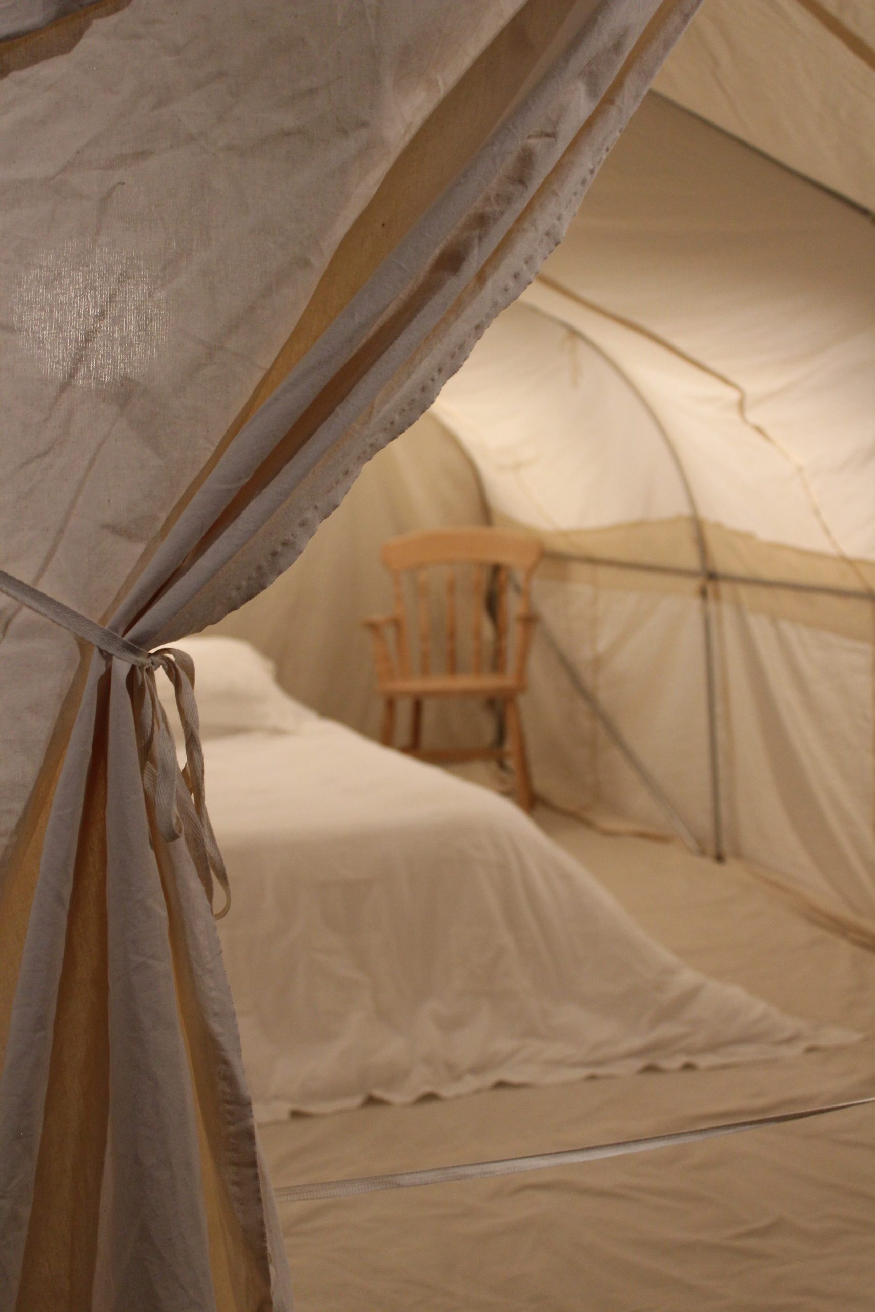 A white curtain pulled back to show the inside of a canvas, cream tent, with a white bed and wooden chair inside.