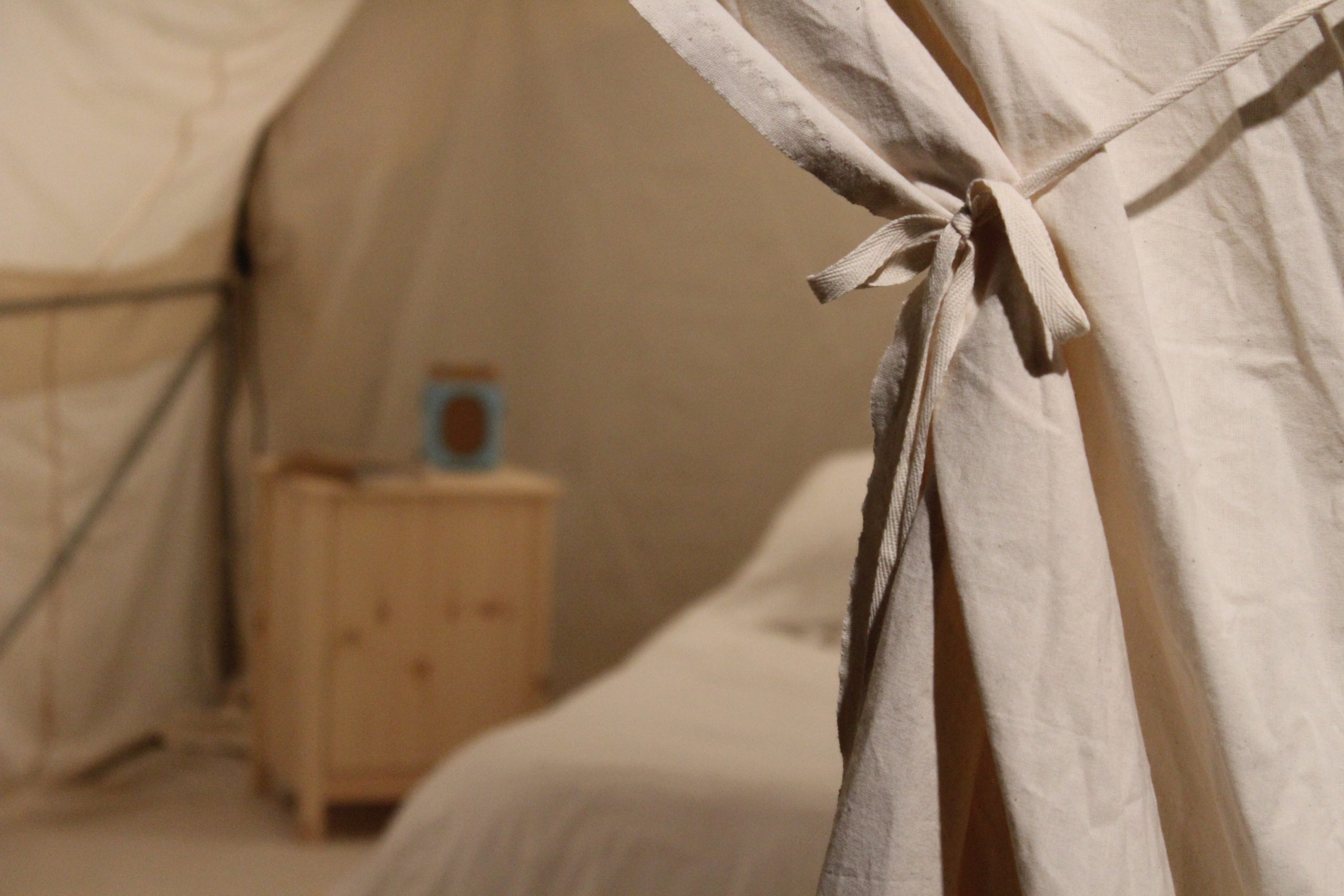 A cream curtain pulled back to show the inside of a tent structure, with a white bed and wooden cabinet.
