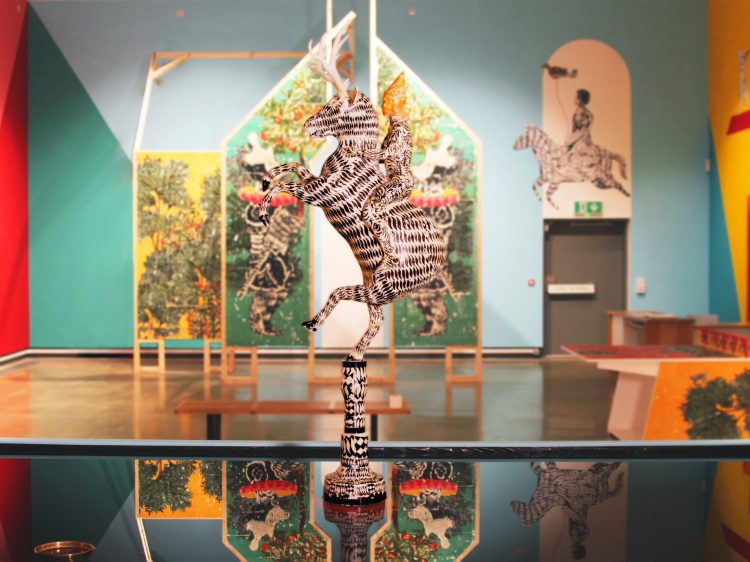 A sculpture of a upright zebra in the middle of a pool of water, with print colourful backgrounds behind it and a wooden house covered in paintings.