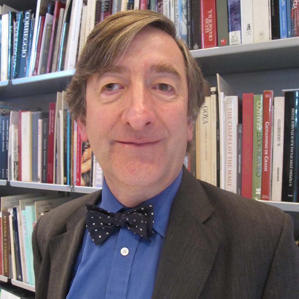 A man with short straight grey-flecked brown hair, wearing a suit and bow tie, standing in front of shelves of books.