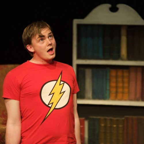 A man looking up at the sky wearing a red shirt with a lightening bolt, with behind him books and bookcases.