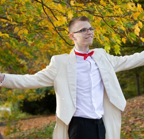 A boy in a white tuxedo and red bowtie, in the forest, conducting music.