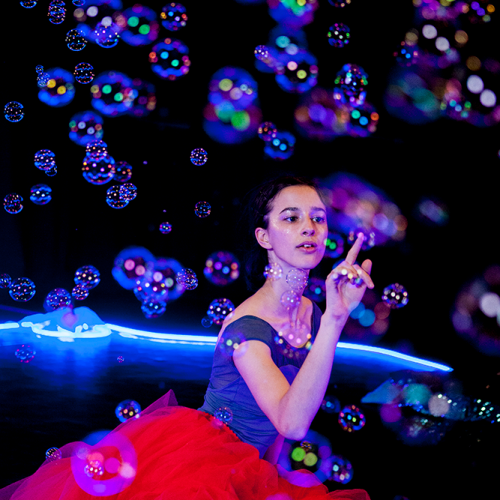 A woman sitting on the floor in a red tutu, surrounded by bubbles in a dark room reaching out to pop one of them.