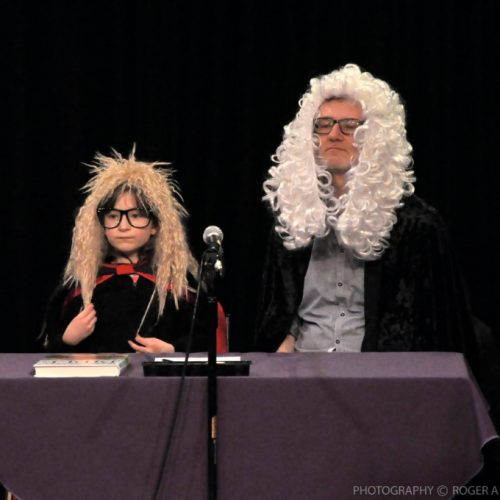 A child and an adult sat at a table, wearing legal wigs and robes in front of a microphone.