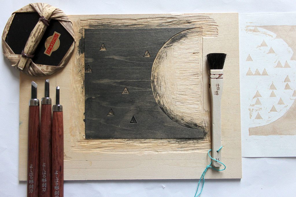 Traditional Mokuhanga tools laying on a wooden base, with a carved moon and triangle print in the middle with ink.
