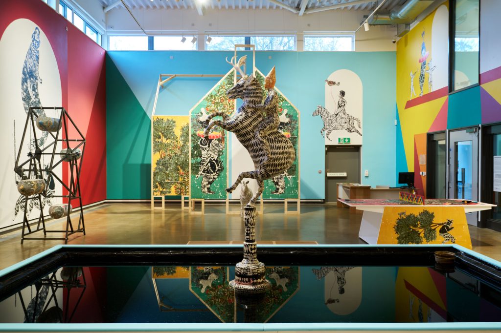 A wide shot of the Mohammad Barrangi exhibition, showcasing a pool of water with a zebra sculpture in the middle, with a wooden painted structure behind it. To the left is a metal Ferris wheel and to the right is a pingpong table.