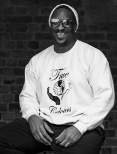 A picture of a man sitting and smiling at the camera, wearing glasses, a light coloured beanie, and a hoodie saying 'True Colours' with a black and a white hand holding the earth.