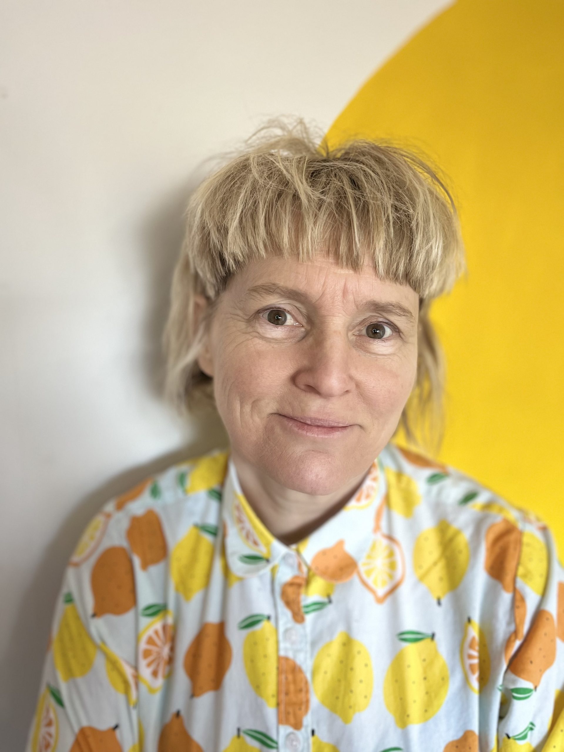 A headshot of a woman smiling at the camera, with short blonde hair. She is wearing a white shirt with illustrations of citrus fruits.