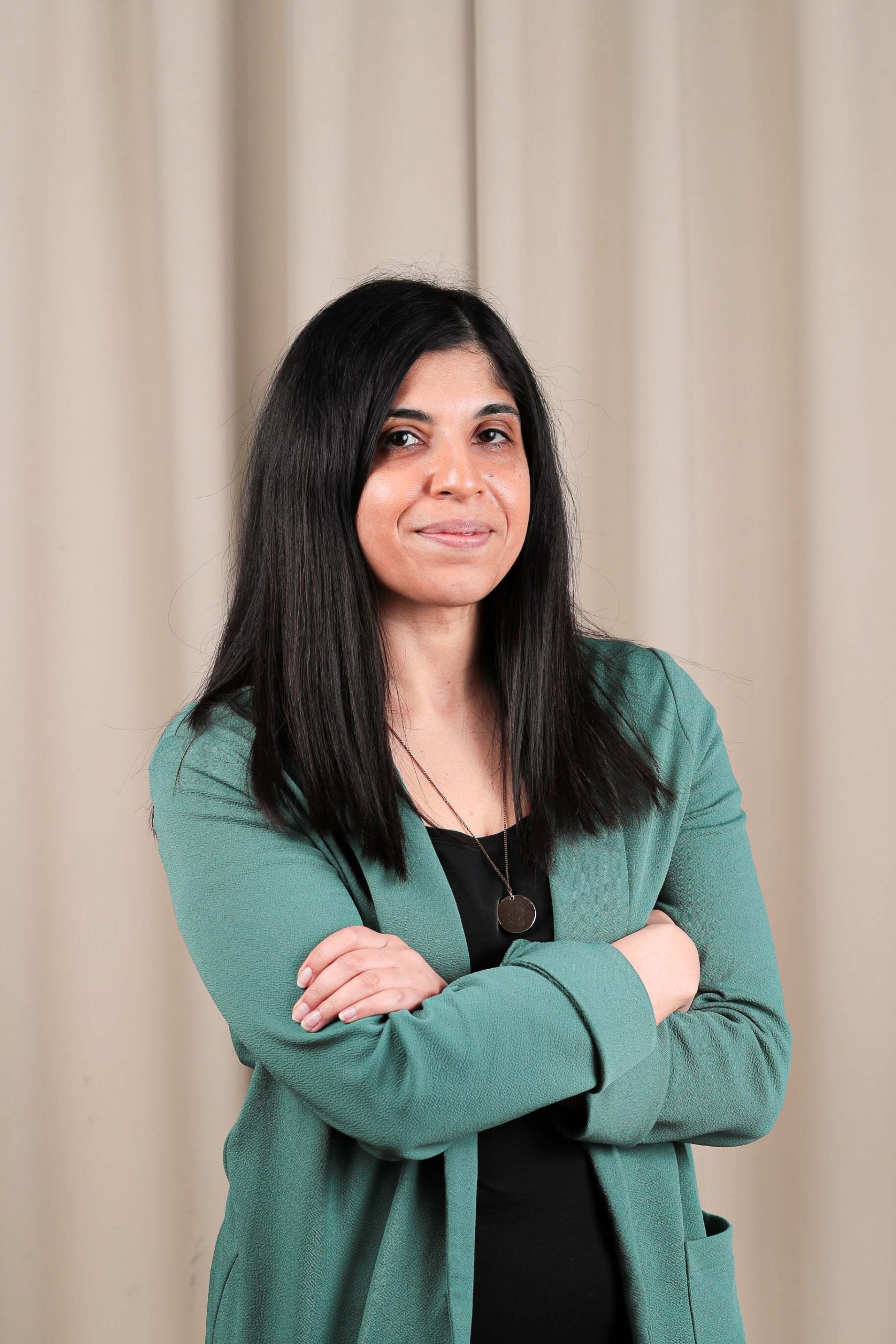 An Asian women with long black hair, arms crossed wearing a teal jacket and black vest top, smiling at the camera.