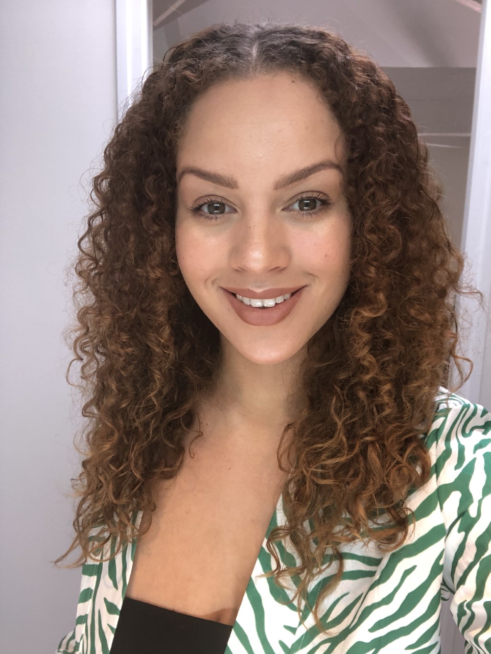 A headshot of a women smiling at the camera, with curly long hair, a black tank-top and zebra green shirt.