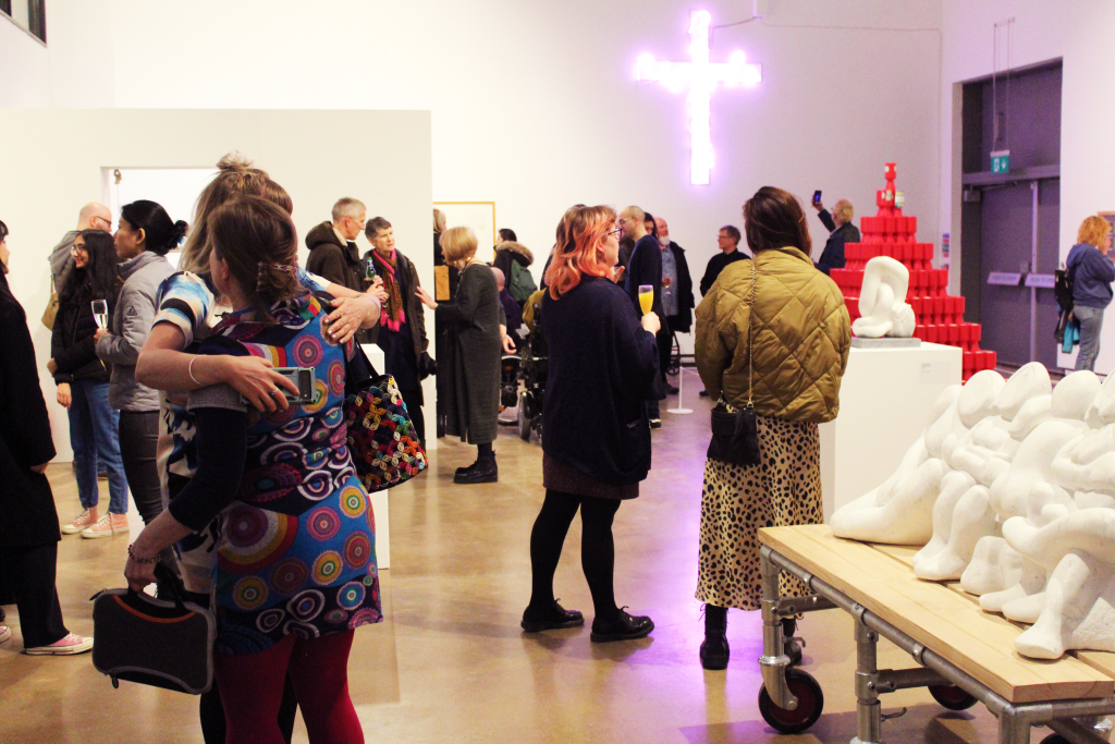 An array of people in Gallery 1 hugging and talking, surrounded by neon lights and marble carvings by Tony Heaton.
