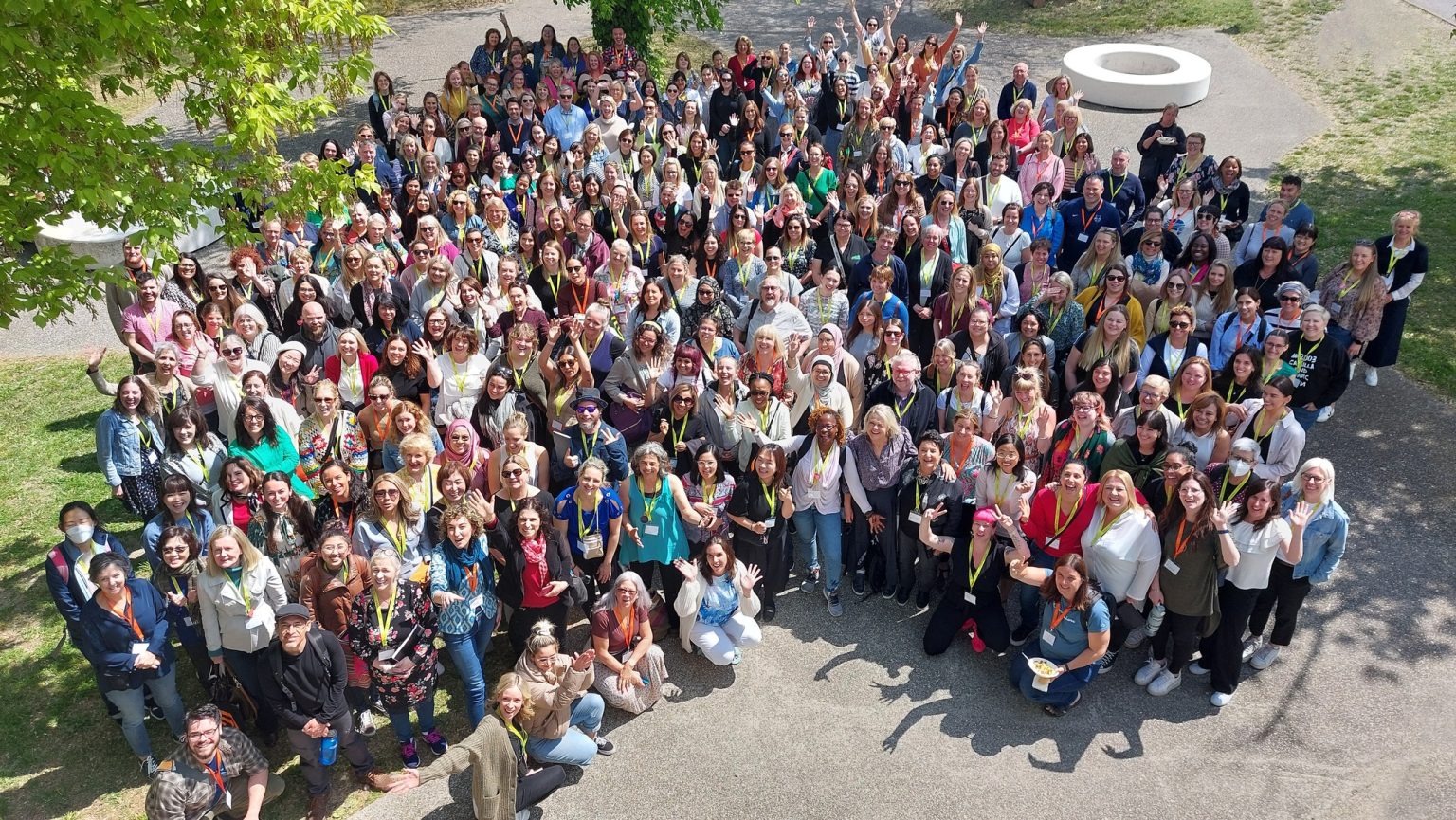An above shot showing a large group of people standing outside in a park, smiling up at a camera.