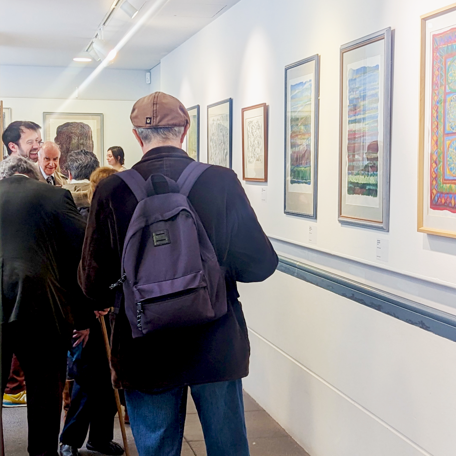 A group of people laughing and smiling in the balcony gallery while they look at Alan Caine's paintings.