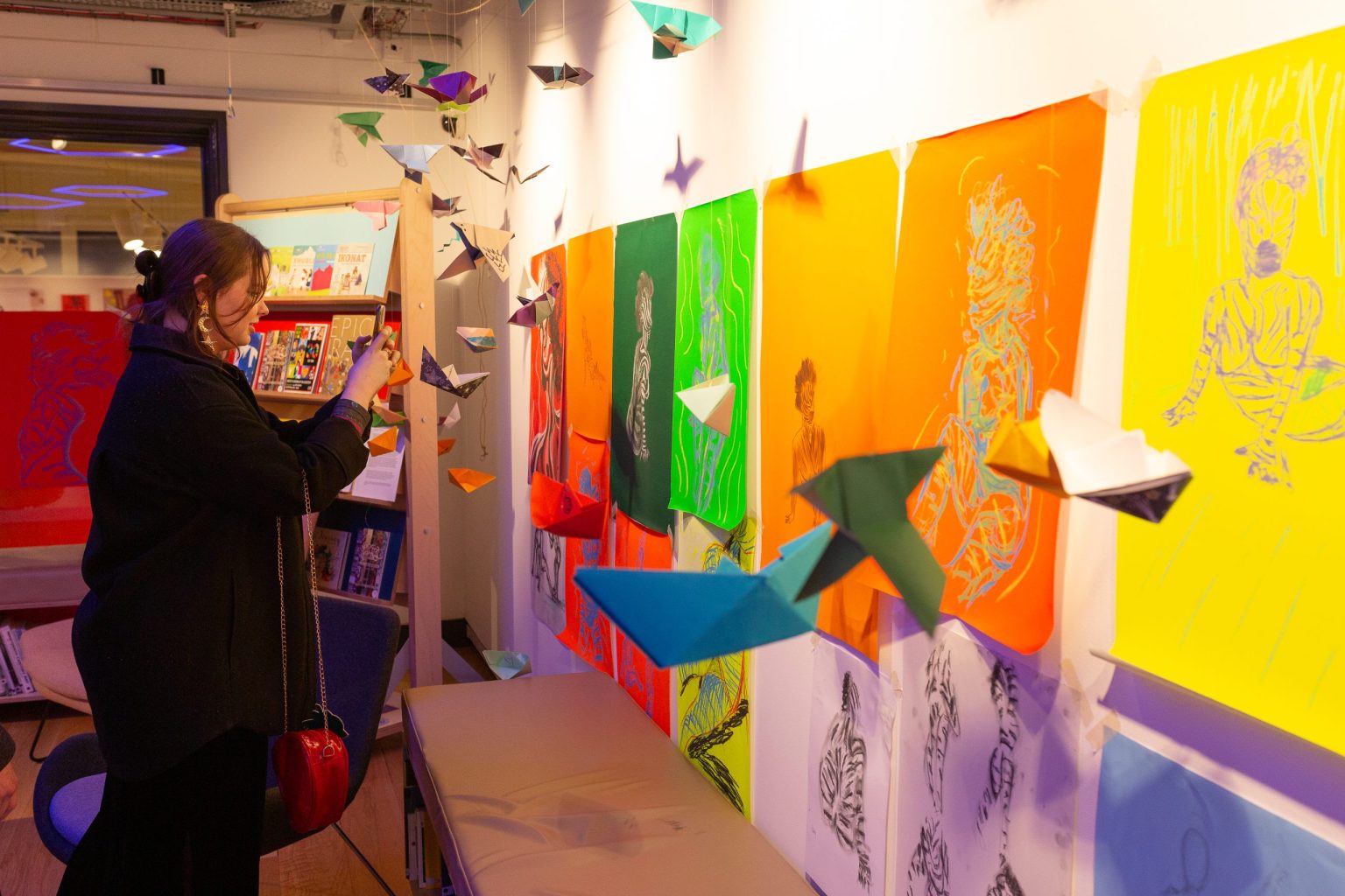 A woman taking photos of origami cranes hanging from the ceiling. On the wall are colourful depictions of a life model painted as a zebra.