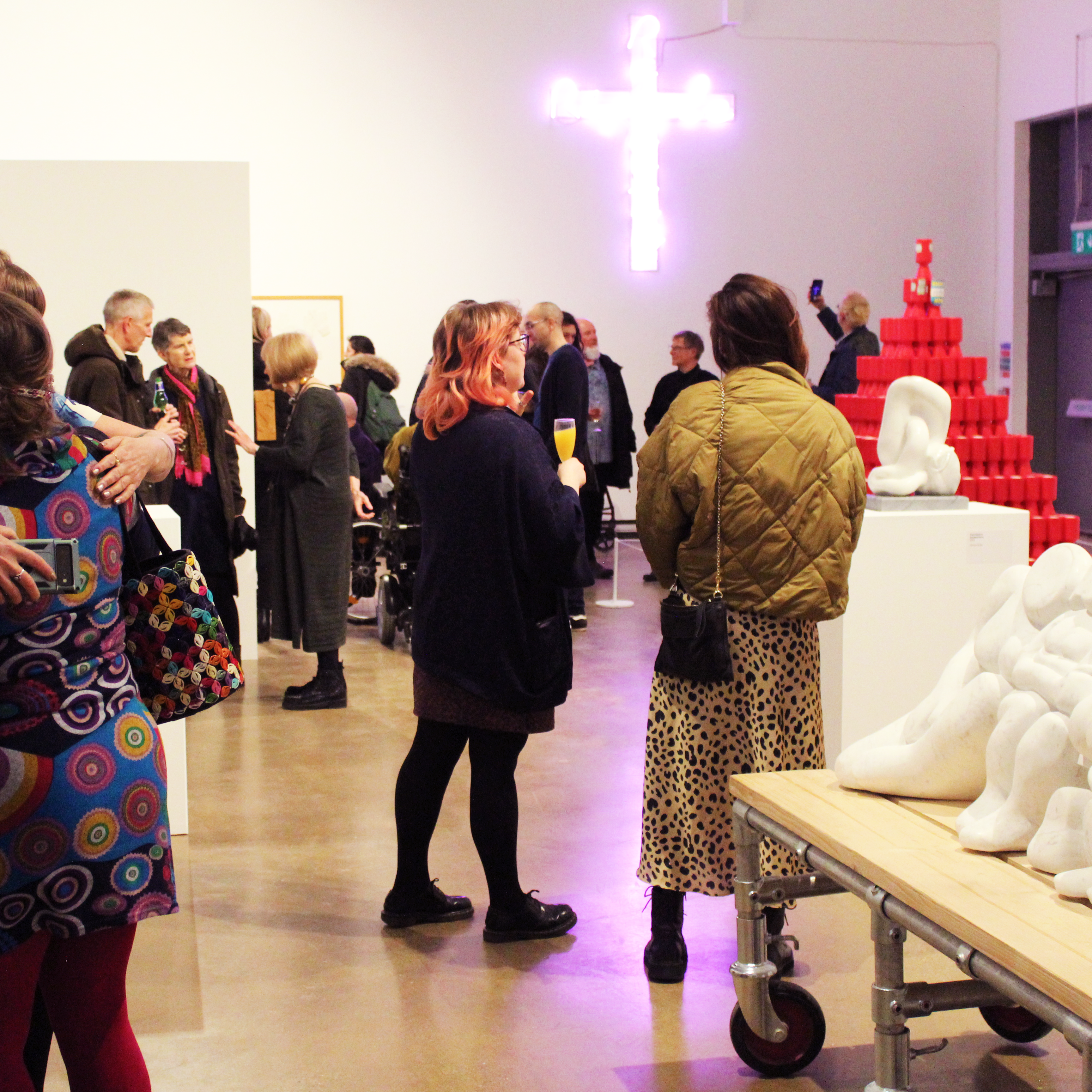 A range of people in Gallery 1 looking at artwork, such as carved marble, neon crosses and pyramids made from red charity boxes.