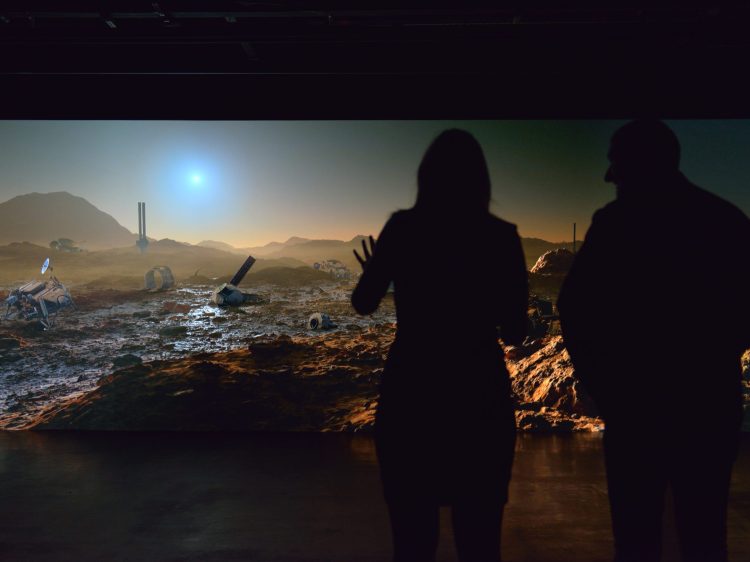 The silhouettes of two people in front of a panoramic view of the surface of Mars.