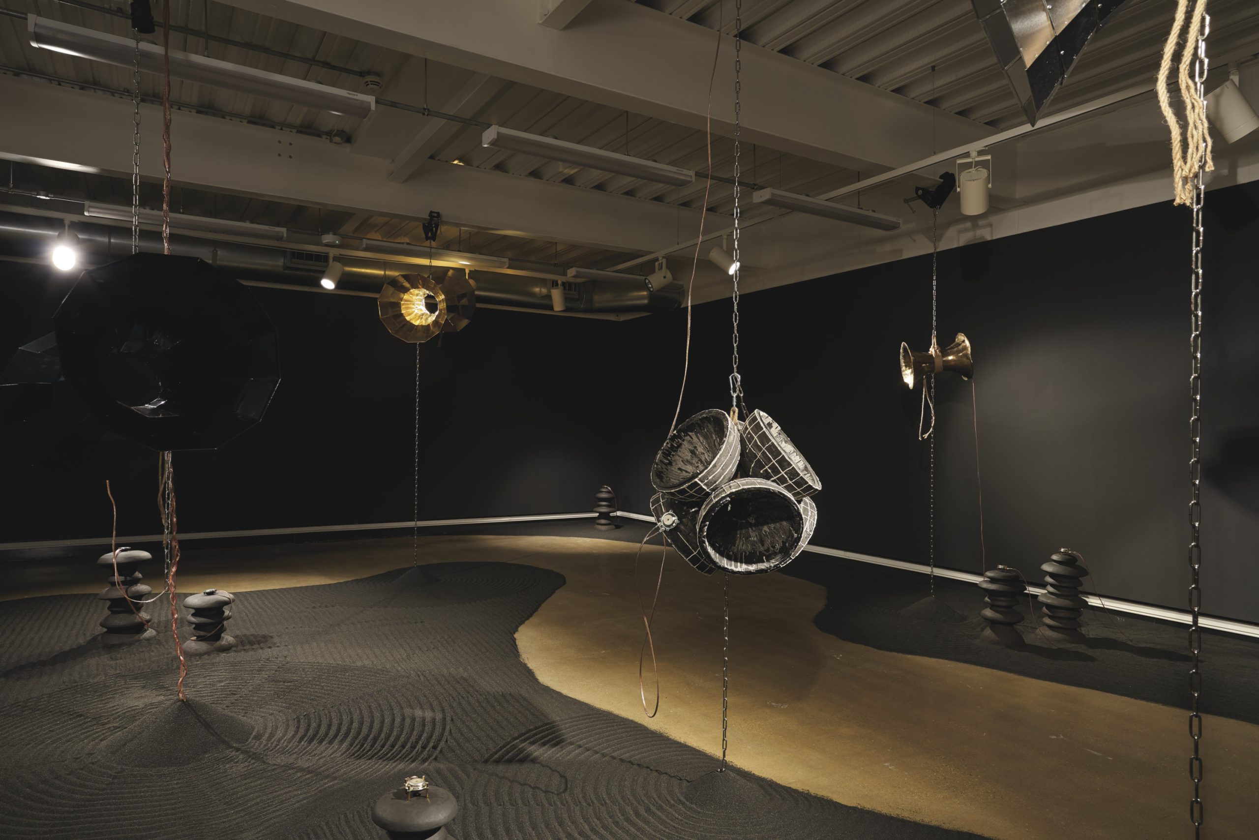 Exhibition space with metal wormholes suspended from the ceiling and black sand in circular patterns with neat towers of rocks on the floor.