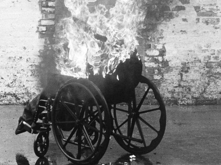 A wheelchair on fire in front of a brick wall in black and white.