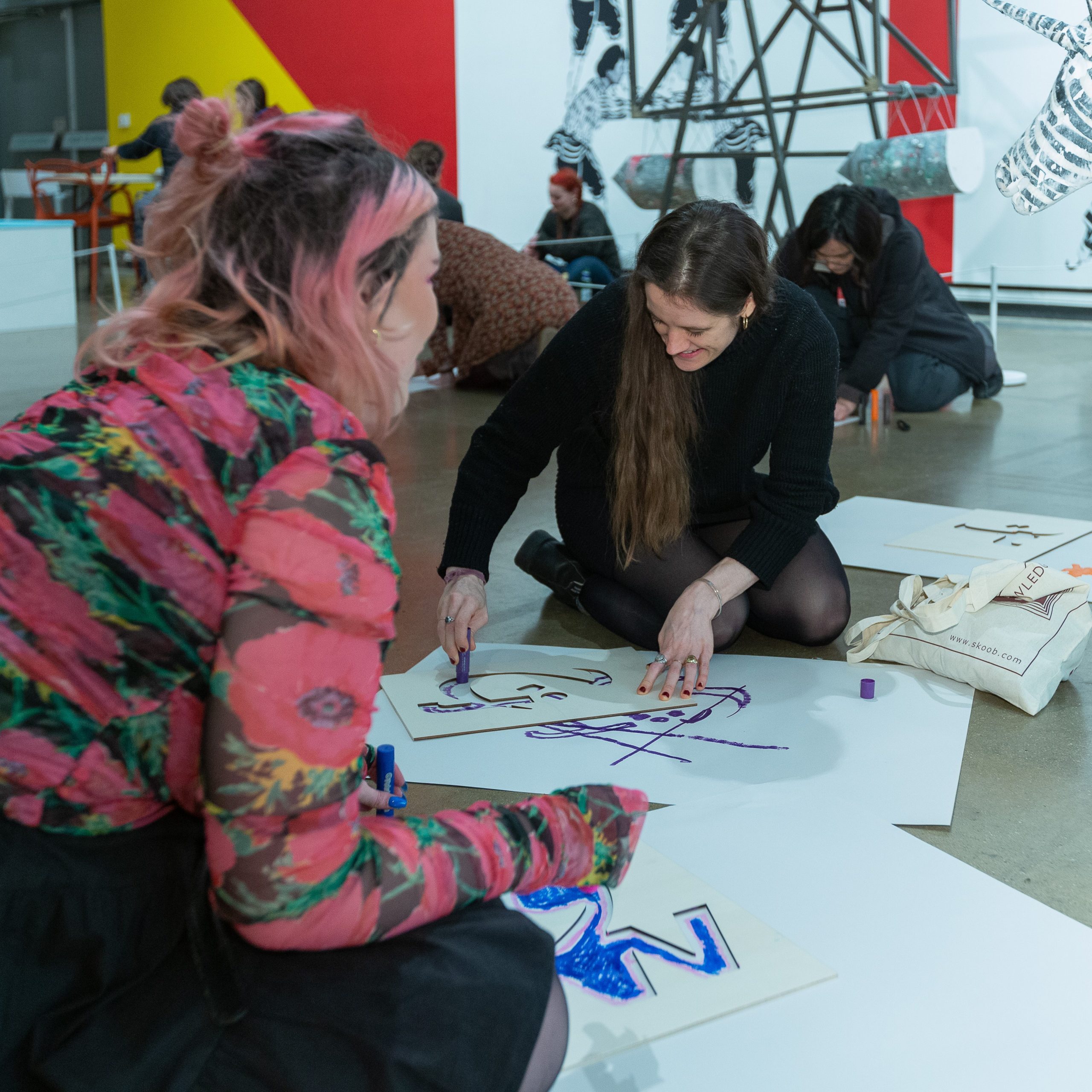 Two white women on the floor of a gallery drawing on pieces of paper.