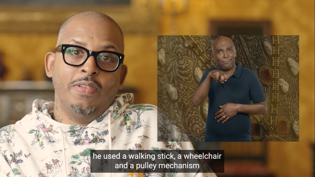 Artist Christopher Samuel talking to the camera while a man next to him interprets his words into BSL. Underneath the caption says "he used a walking stick, a wheelchair and a pulley mechanism."