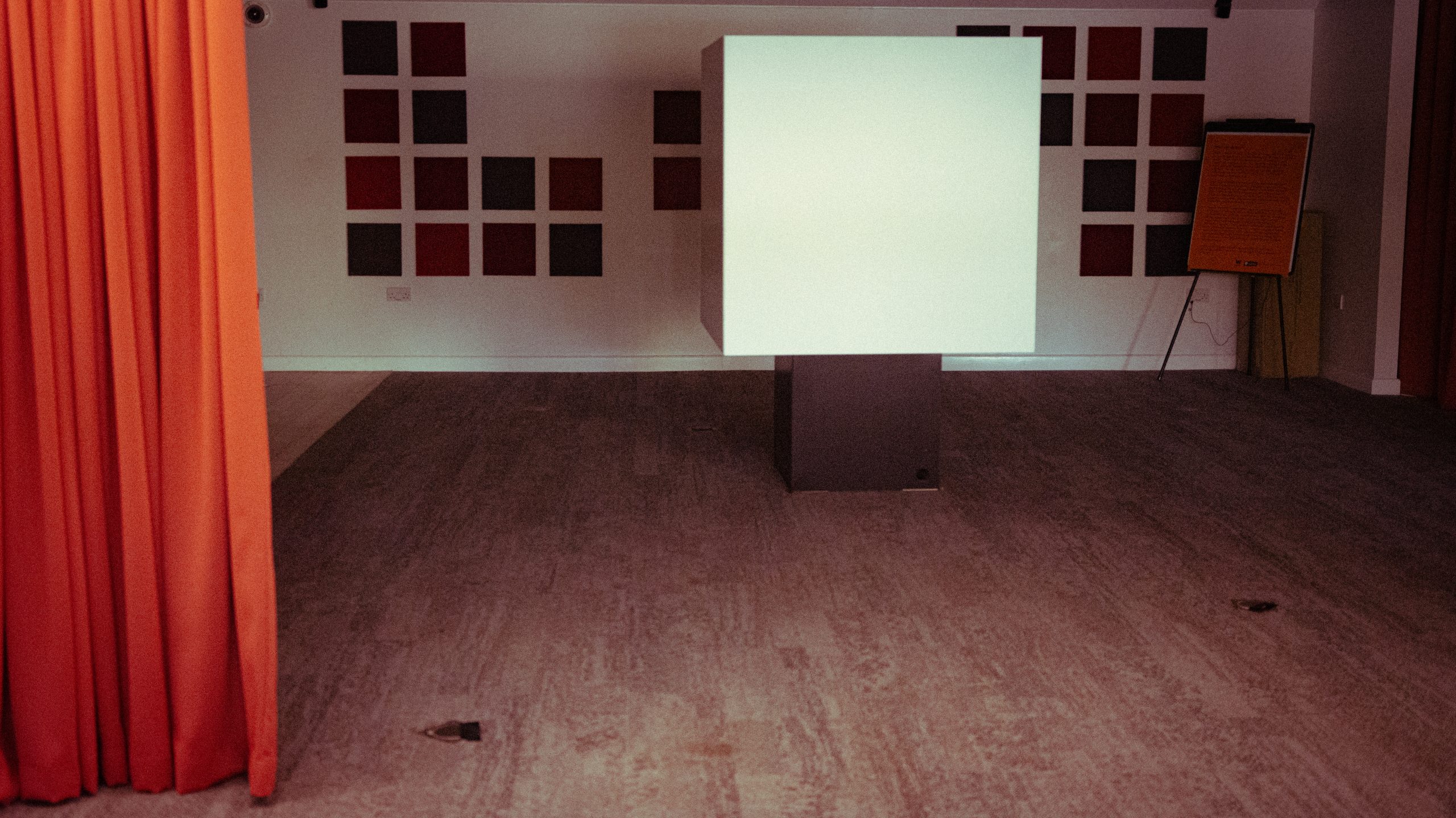 A dark room with an orange curtain to the side. In the middle stands a large white cube on a black stand.