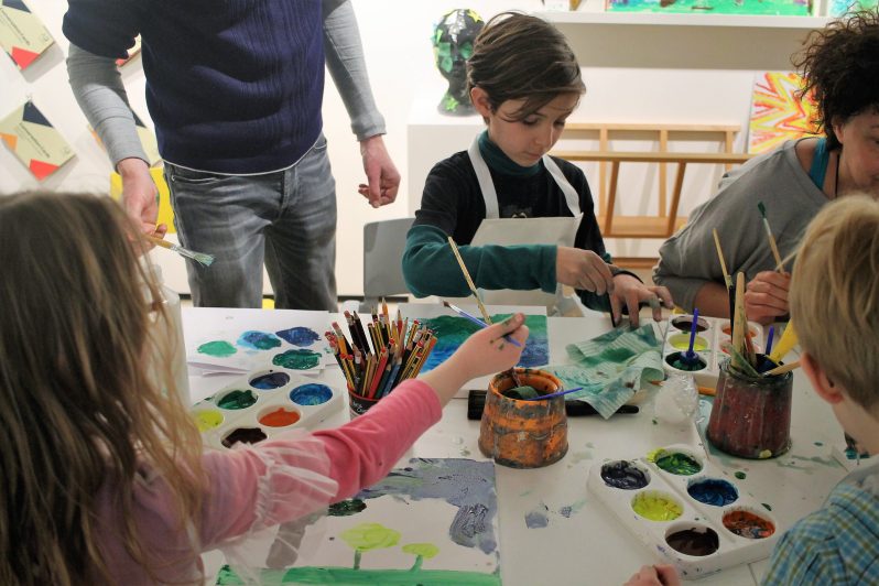 Children and adults around a table covered in paints and paintbrushes, making art on paper.