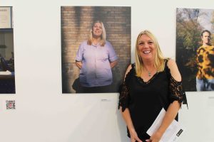 A white women with blonde hair and a black shirt smiling next to a portrait of herself on the wall.