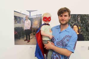 A white man with a blue shirt holding his child, who is wearing a red and blue cape and red superhero mask. They are smiling next to a portrait of the man.