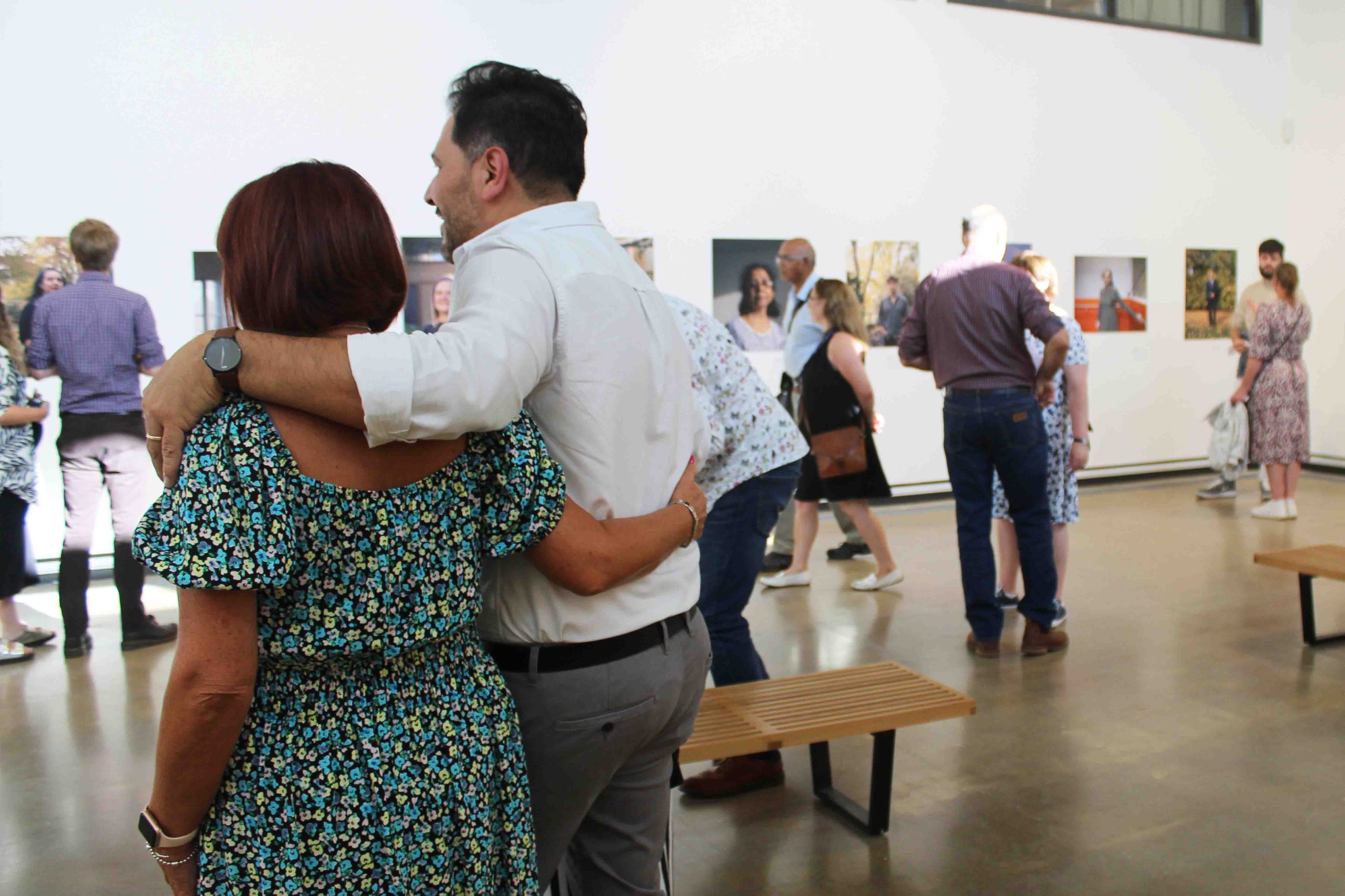 A man and a women with their arms around each other and their backs to the camera. Beyond them are people in a gallery.