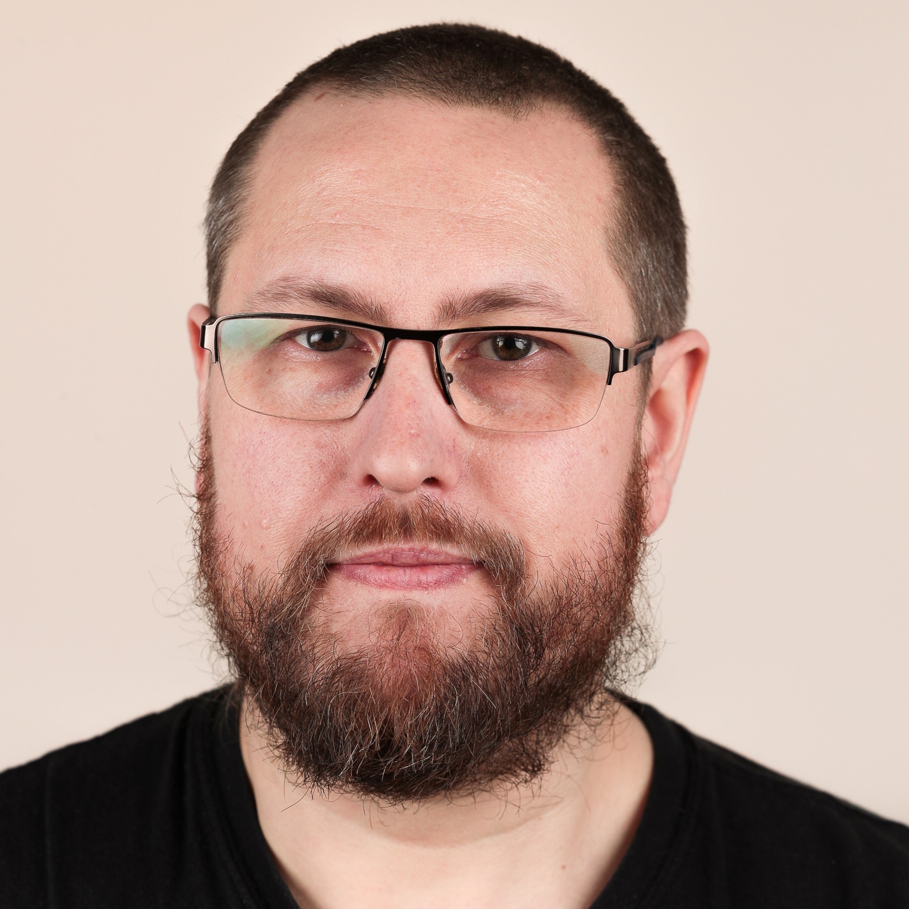 A man with a dark buzzcut and a dark beard flecked with grey, wearing rectangular glasses, looks into the camera.