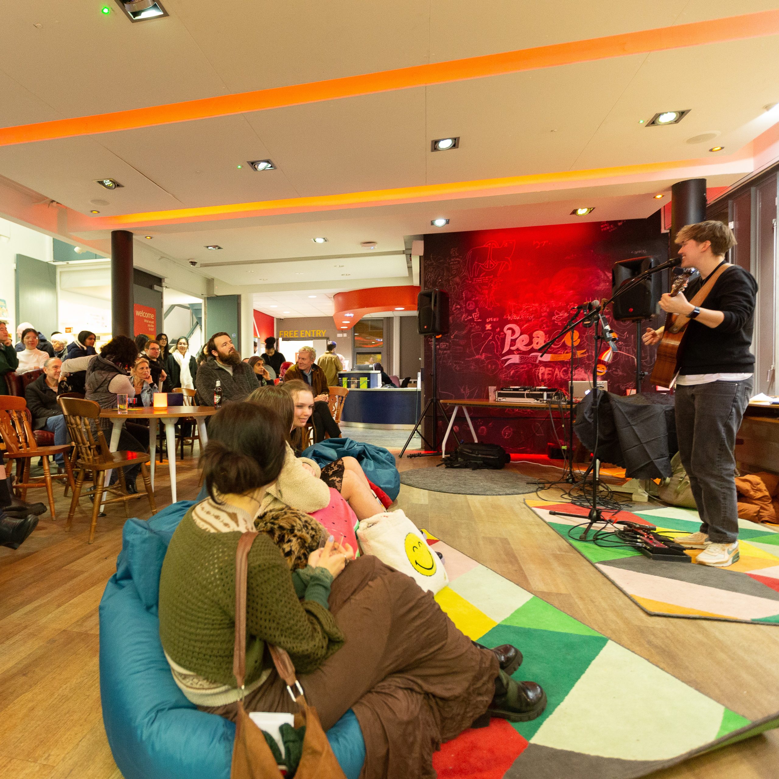 A group of people sitting on chairs or bean bags on the floor to the left, watching a person with a guitar and microphone perform music to the right.