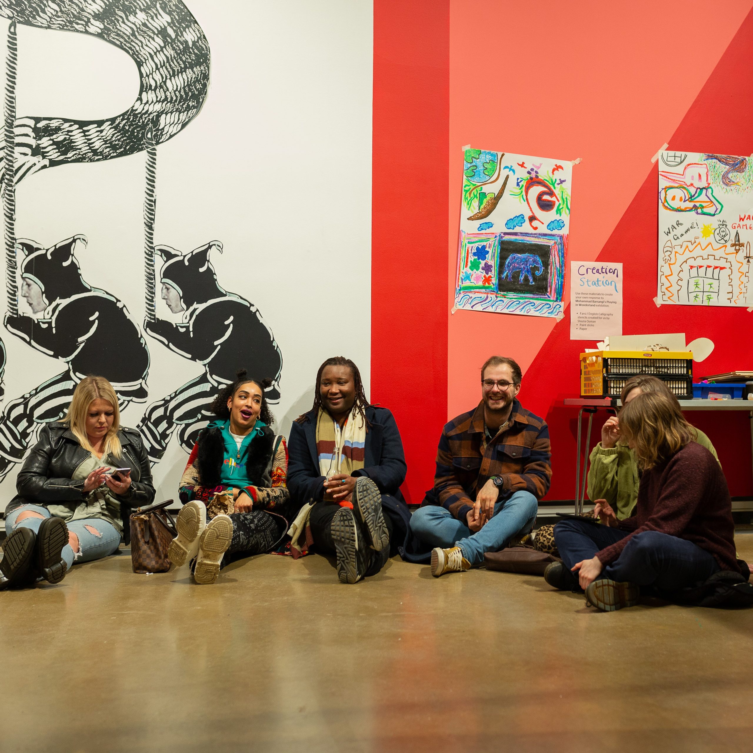 A group of five people sat on the floor of a gallery talking and laughing together. They are leaning against colour painted walls, with self-made crayon artwork stuck onto the wall.