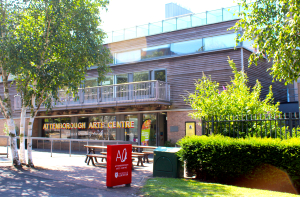 The outside of Attenborough Arts Centre building on a sunny day surrounded by green trees and nature. On the windows the name of the centre is painted in white, red and yellow signwriting.